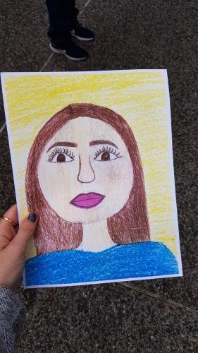 "Mask" - a self portrait drawn by a child. A young girl with brown hair and a blue shirt on a yellow background.