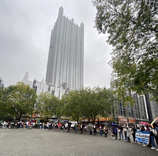 Local university students, community members and HCEF staff gathered in Market Square, Downtown Pittsburgh to represent the homeless youth identified in Allegheny County. It is an overcast day with a light mist. The ground is wet and people are in jackets and have umbrellas.